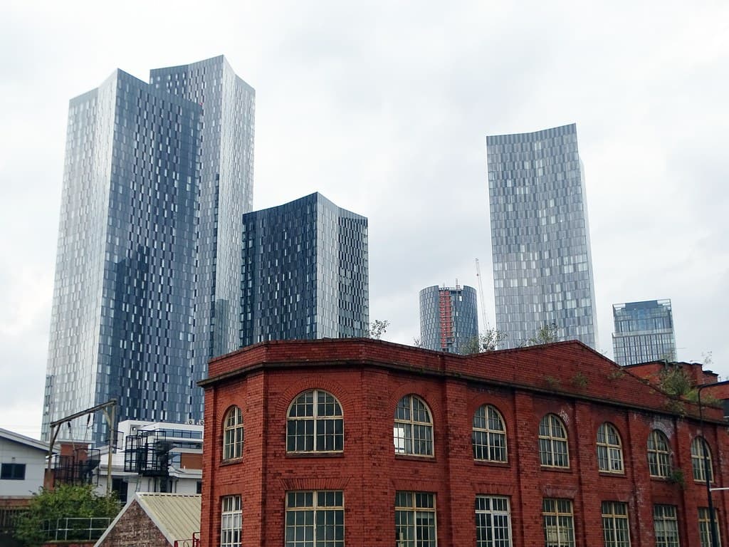 Cluster of tall modern glass residential towers rising behind a red-brick Victorian industrial building in central Manchester.