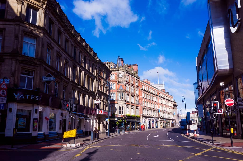 A street in Leeds city centre lined with period and modern commercial buildings under a clear blue sky.
