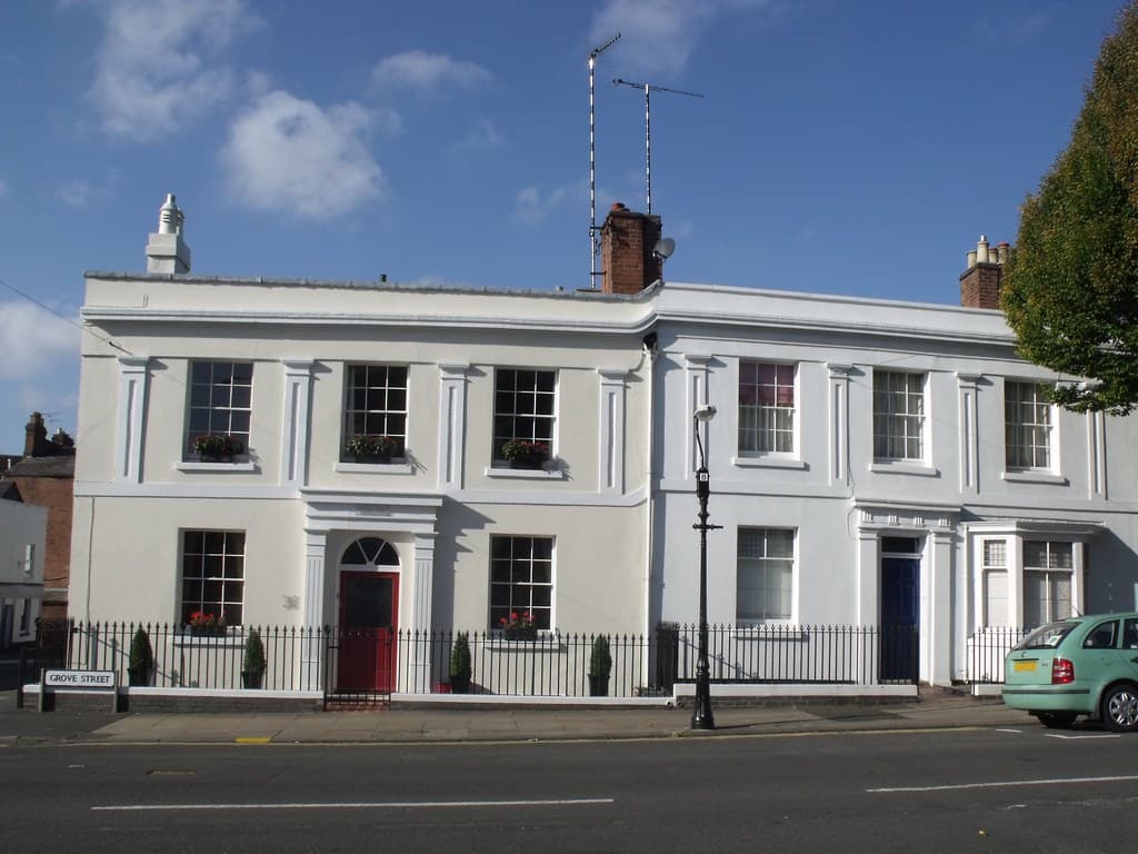 A terrace of white-rendered Georgian houses with red and blue front doors, iron railings and a Grove Street sign, on a residential street in Leamington Spa, England.