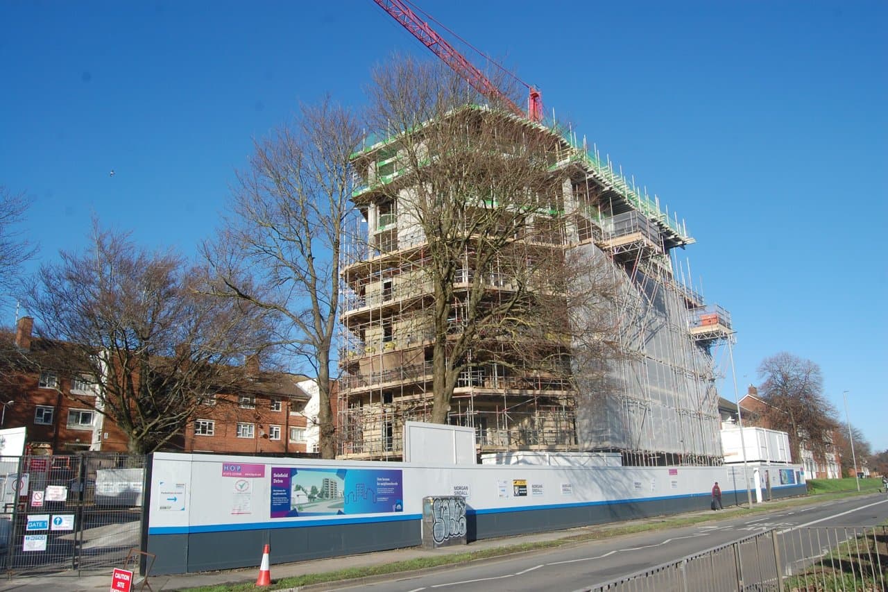 An eight-storey block of housing association flats under construction in Brighton, England, clad in scaffolding with a red tower crane against a clear blue sky.